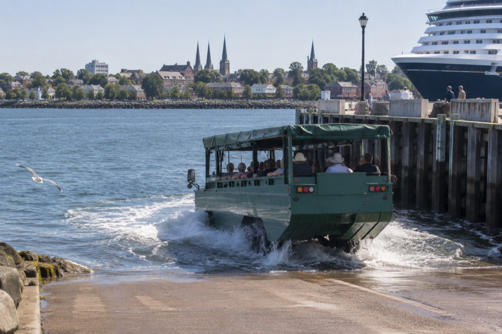 An amphibious Harbour Hopper vehicle driving down a ramp into the water, with passengers waving from the open sides.