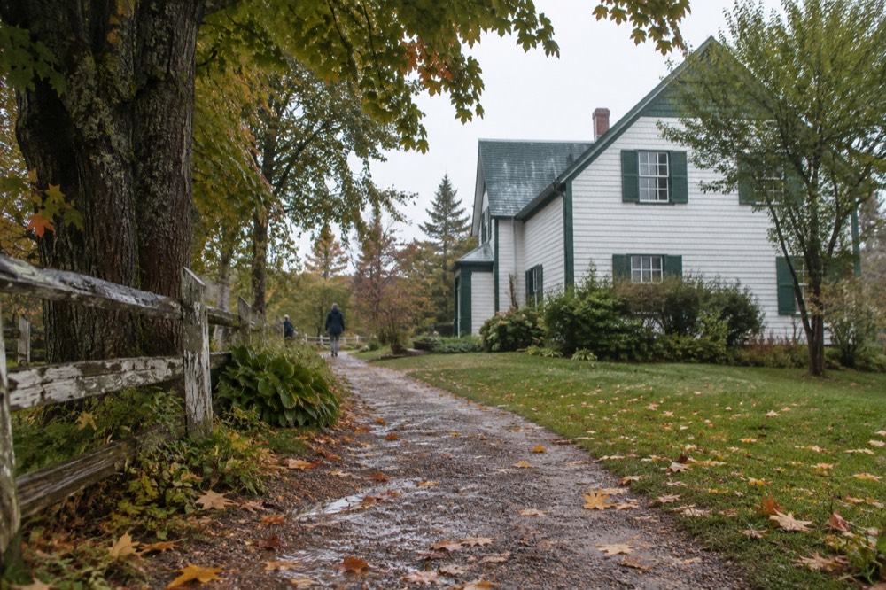 Wooden farmhouse with green trim beside a garden path under tall trees