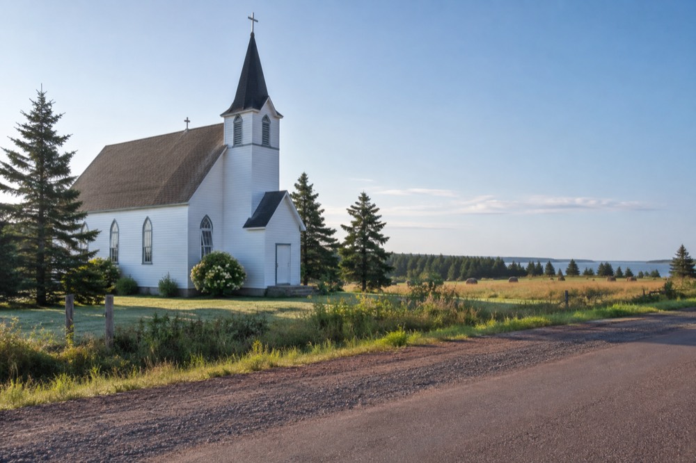 A small white church beside a quiet rural road with green fields behind it