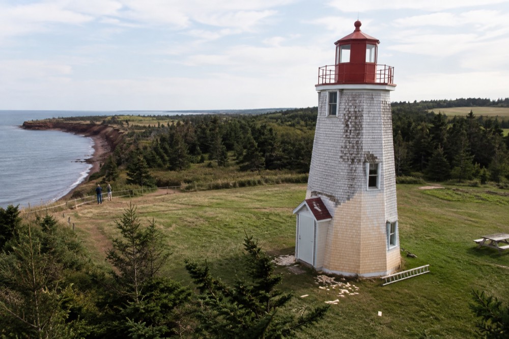 A round white lighthouse with a red lantern room standing on a grassy point above red cliffs