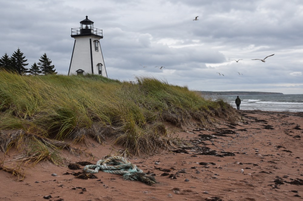 A white lighthouse with black trim standing on a sandy point with dunes and calm water behind it