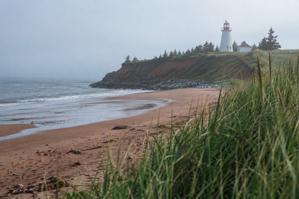 A lighthouse seen across a red sand beach with dune grass in the foreground