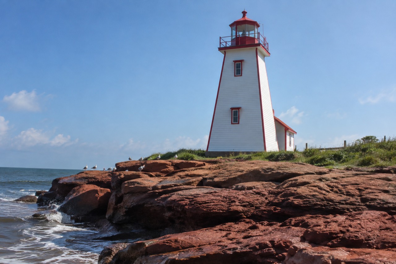 A tall white lighthouse with red trim standing on a rocky point above red sandstone cliffs and open water