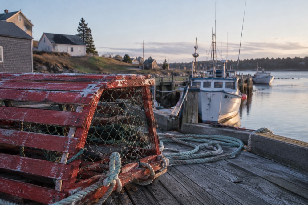 A red lobster trap stacked on a wooden wharf beside fishing boats
