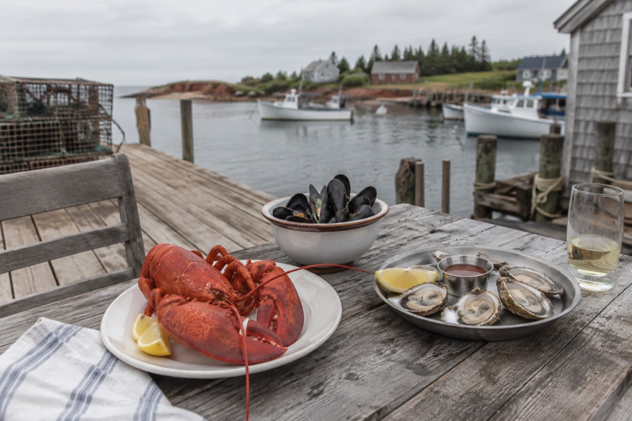 A wooden dock table set with a plate of lobster, a bowl of mussels and a tray of oysters beside a harbour