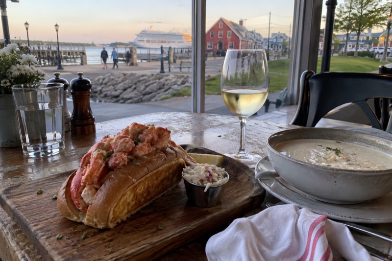 A lobster roll on a wooden board beside a bowl of chowder and a glass of white wine on a Charlottetown restaurant table