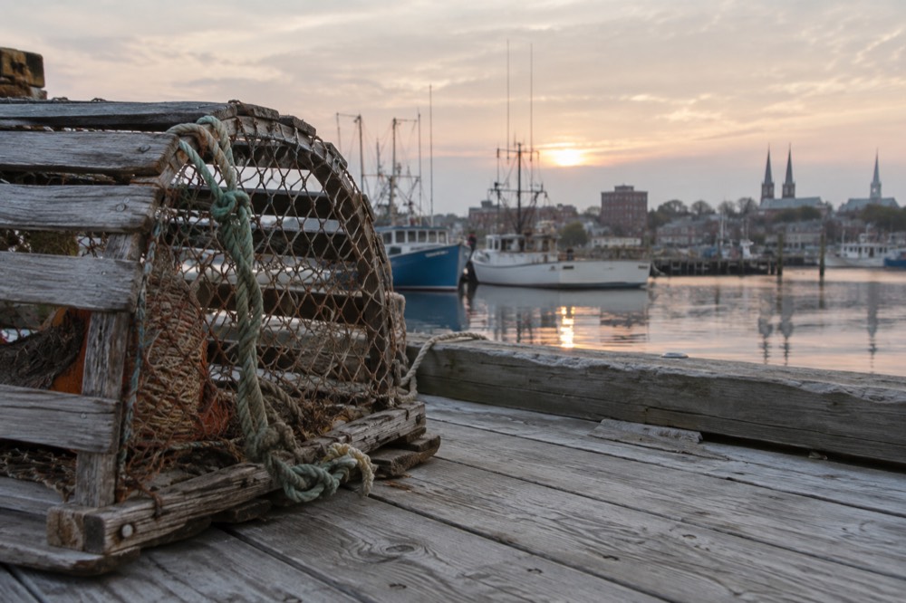 A wooden lobster trap stacked on a red wharf beside fishing boats