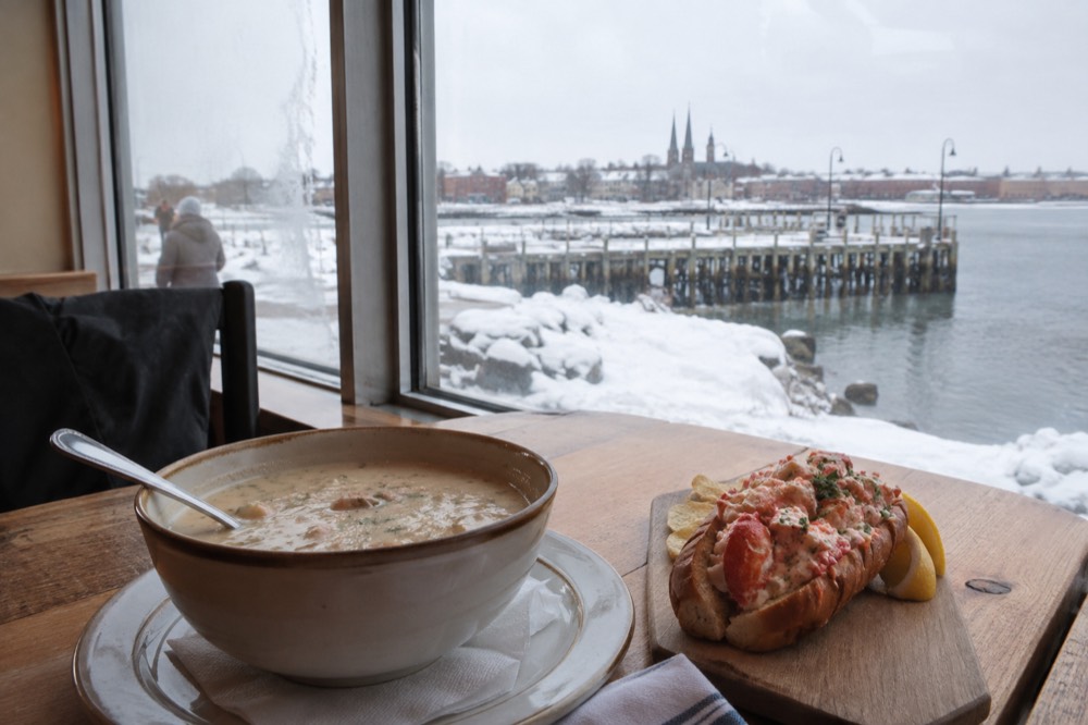 A bowl of chowder and a lobster roll served on a wooden table beside a window