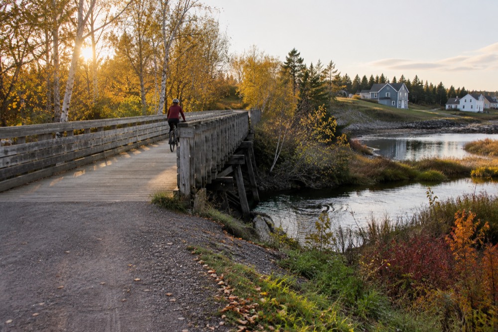 A cyclist on a flat stone dust trail crossing a wooden bridge over a stream