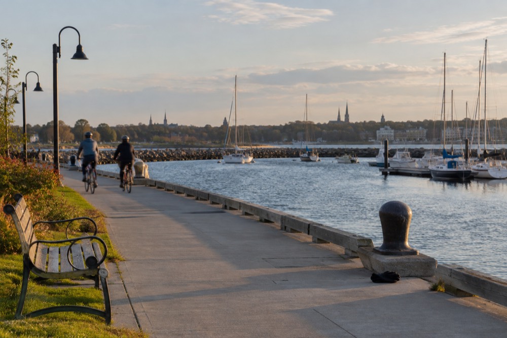 Cyclists riding an e-bike along a waterside path with sailboats moored nearby