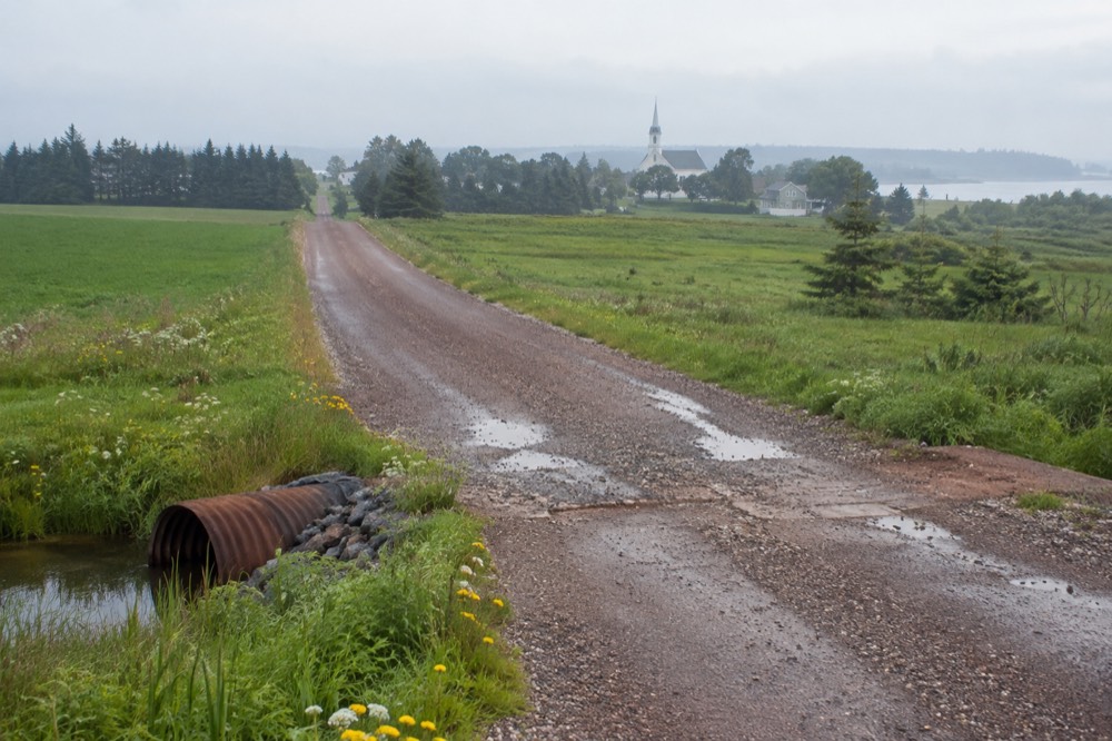 A long straight stone dust trail cutting through green fields toward a distant church spire