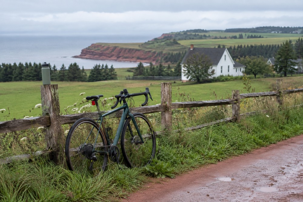 A bicycle leaning against a wooden fence with farmland and a farmhouse behind it